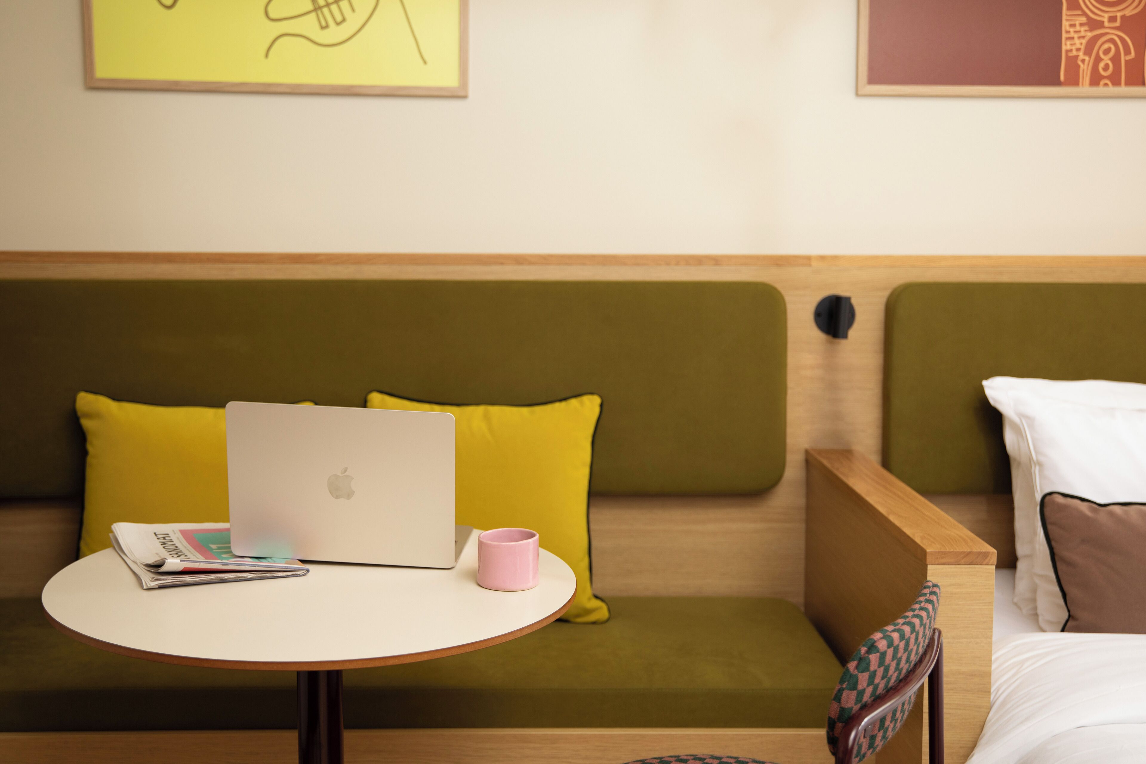 A modern hotel room with a green sofa, a wooden side table with a laptop and a pink mug, and a bed with white and brown pillows.