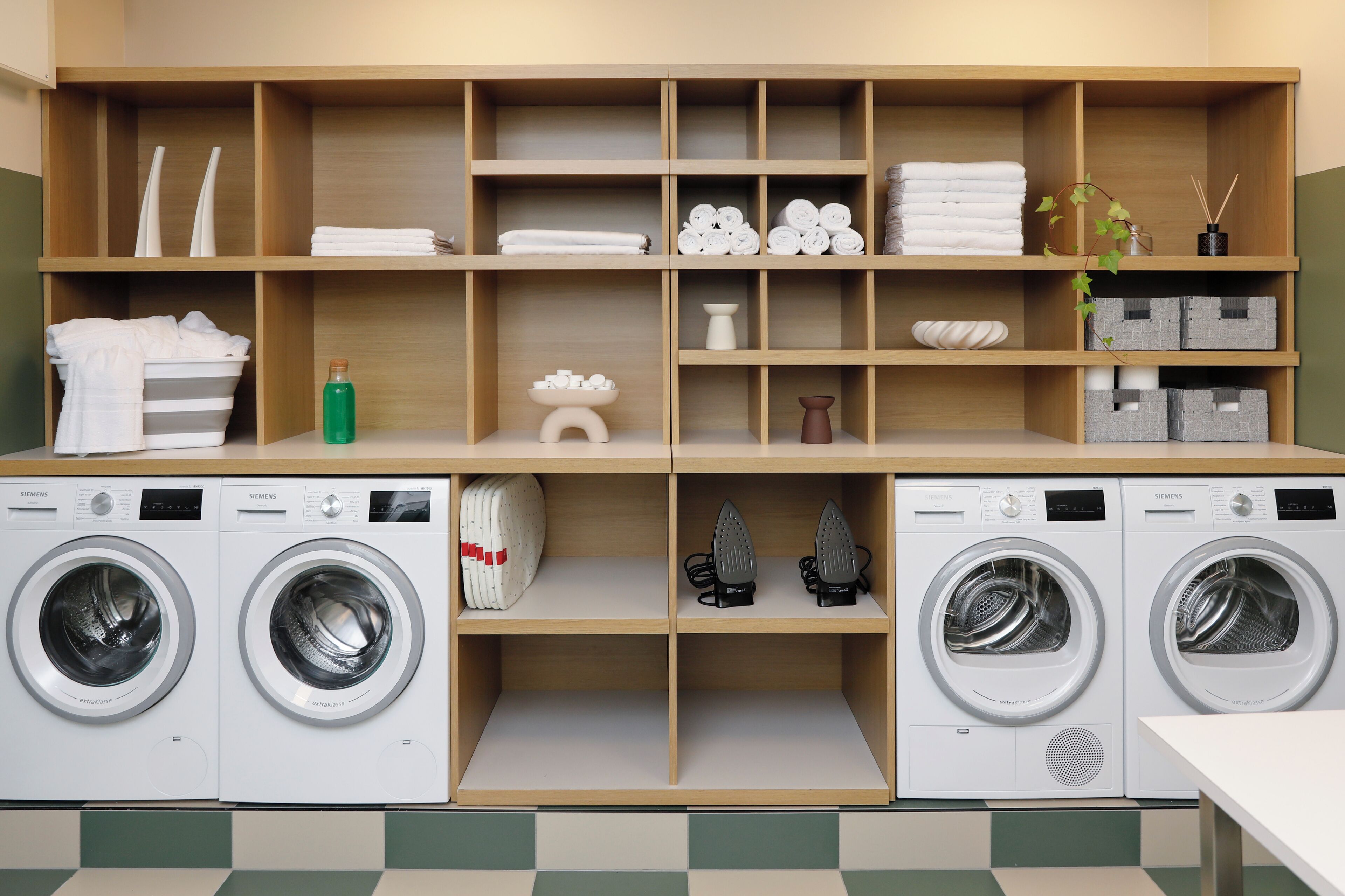 A laundry room with two white Siemens washing machines, wooden shelves with towels, toiletries, and a plant.