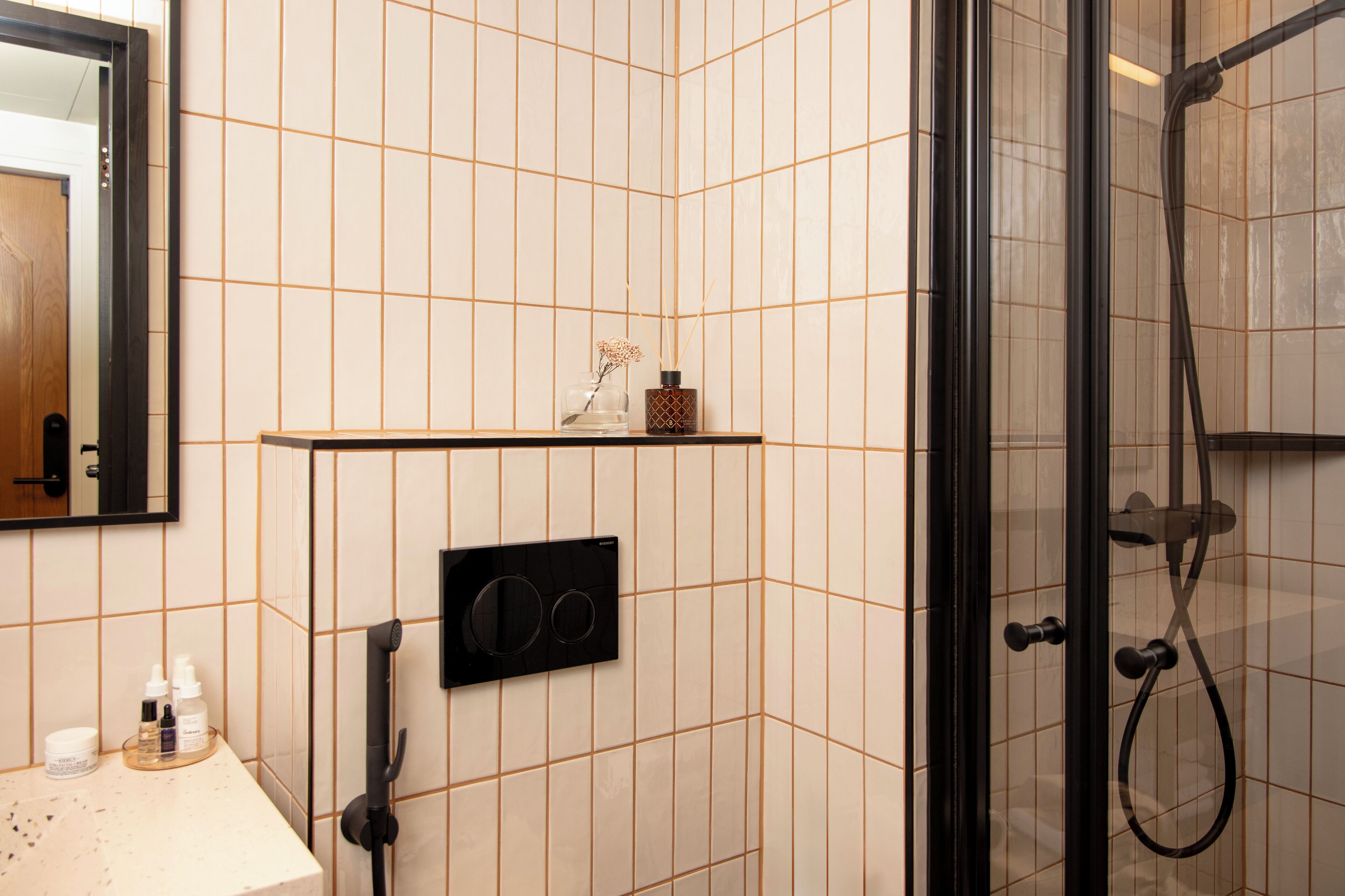 A bathroom with a tiled wall, a mirror, a shelf with toiletries, and a shower area.