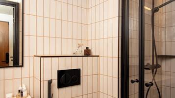 A bathroom with a tiled wall, a mirror, a shelf with toiletries, and a shower area.