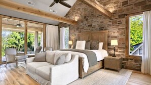 A bedroom with a stone wall, a large bed, a wooden ceiling fan, and a view of greenery outside.