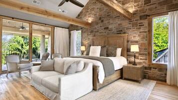 A bedroom with a stone wall, a large bed, a wooden ceiling fan, and a view of greenery outside.
