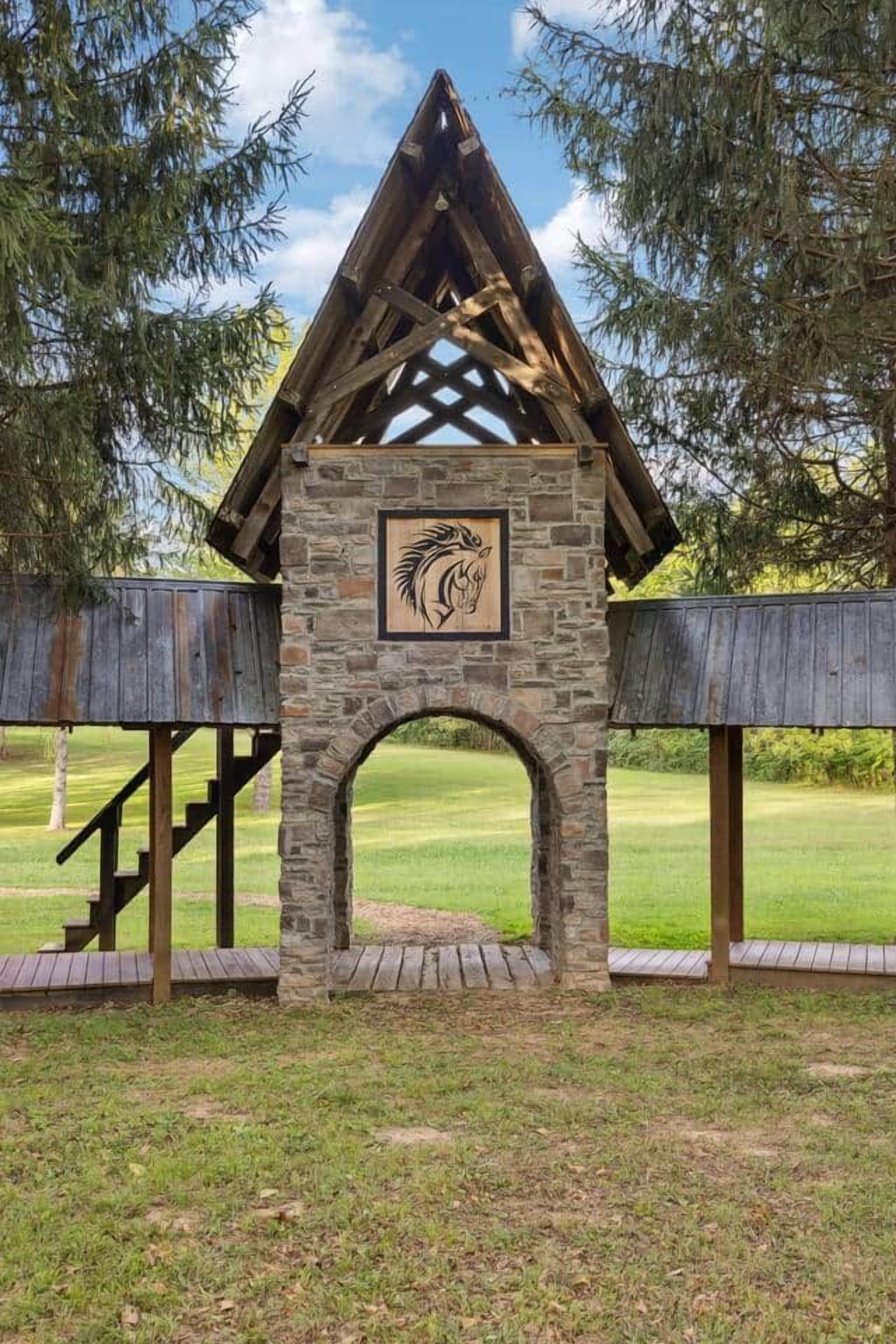 Une structure en pierre surmontée d’un toit en bois et ornée d’un logo, entourée d’une zone herbeuse et d’arbres.