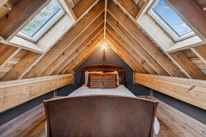 A loft bedroom with a sloped ceiling, wooden beams, a bed with pillows, and skylights.