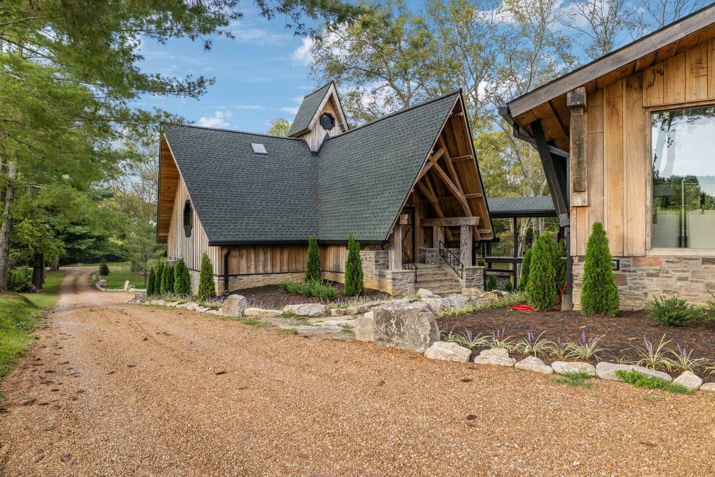 A rustic cabin with a steeply pitched roof, stone foundation, and a gravel driveway leading to a wooden entrance.