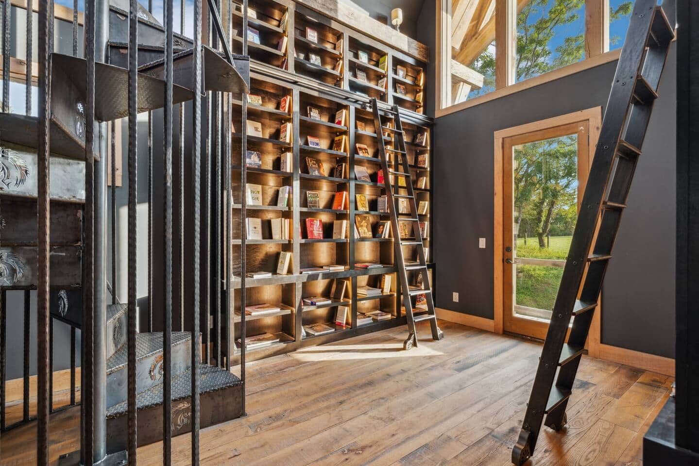 Una habitación con una amplia estantería llena de libros, una escalera de madera y vistas al exterior a través de un ventanal.