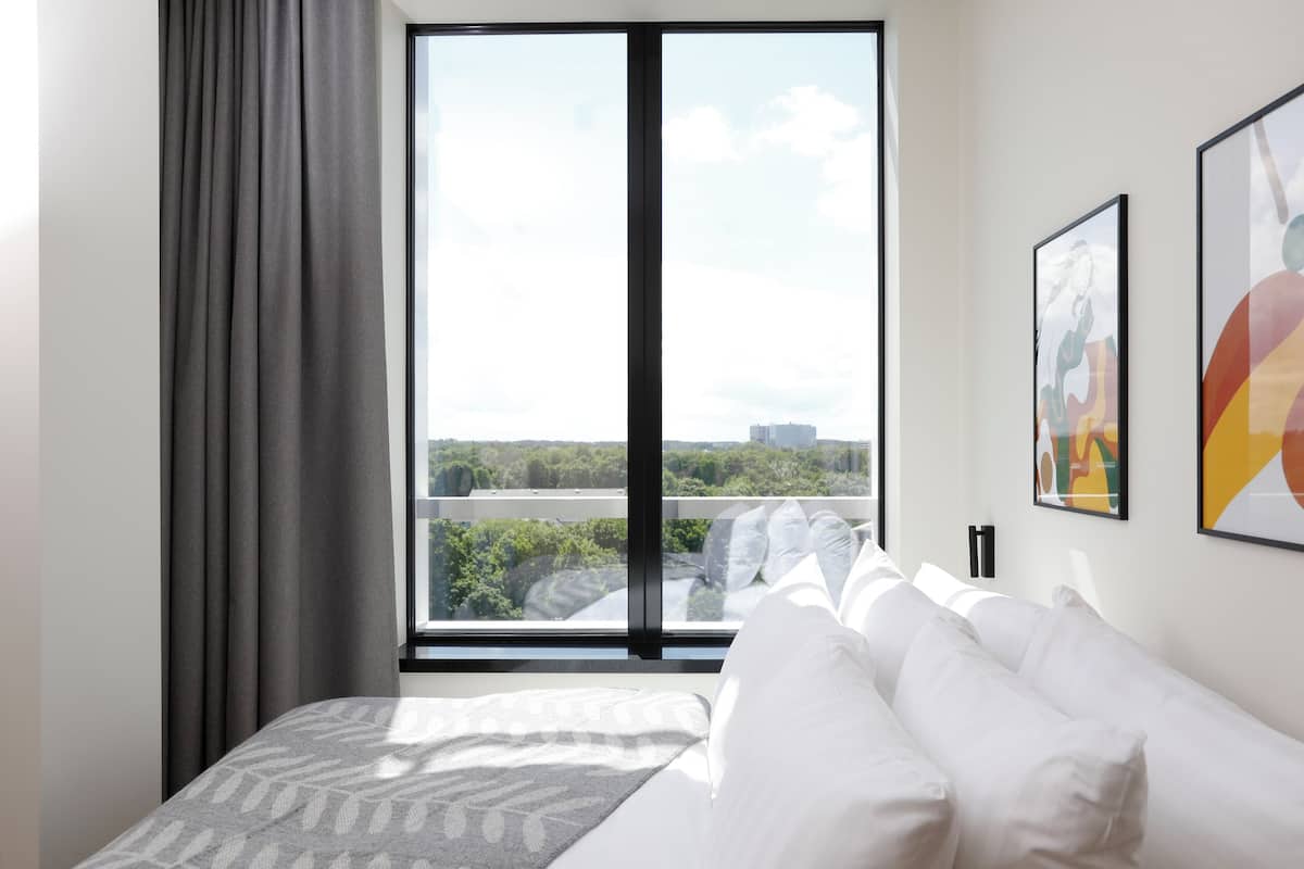 Una habitación de hotel moderna con un ventanal grande, una cama con ropa blanca y dos cuadros abstractos en la pared.