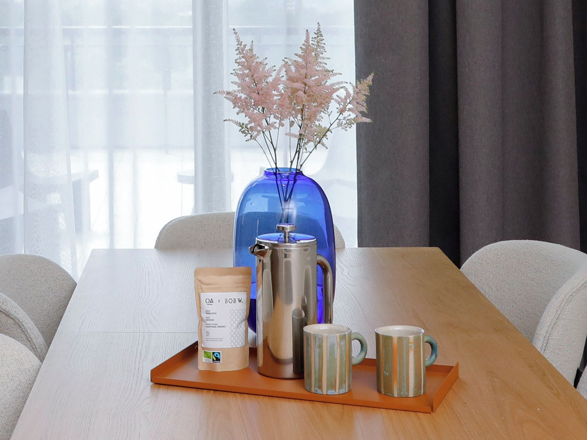 A wooden table with a coffee pot, two mugs, a bottle of coffee, and a blue vase with flowers.