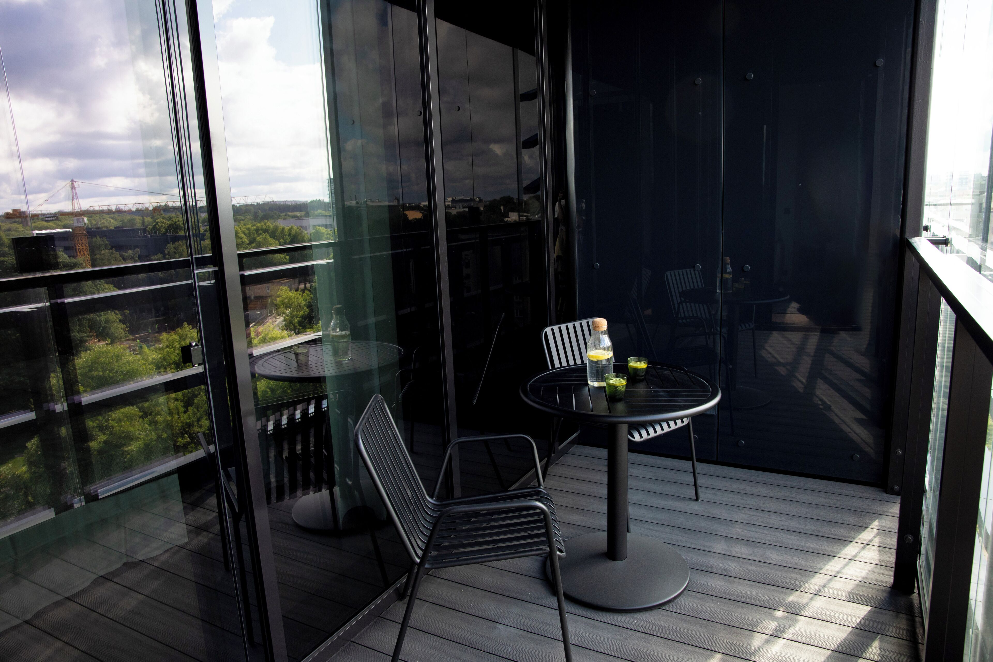A balcony with a glass wall, a round table, two chairs, and a view of greenery and a distant structure.