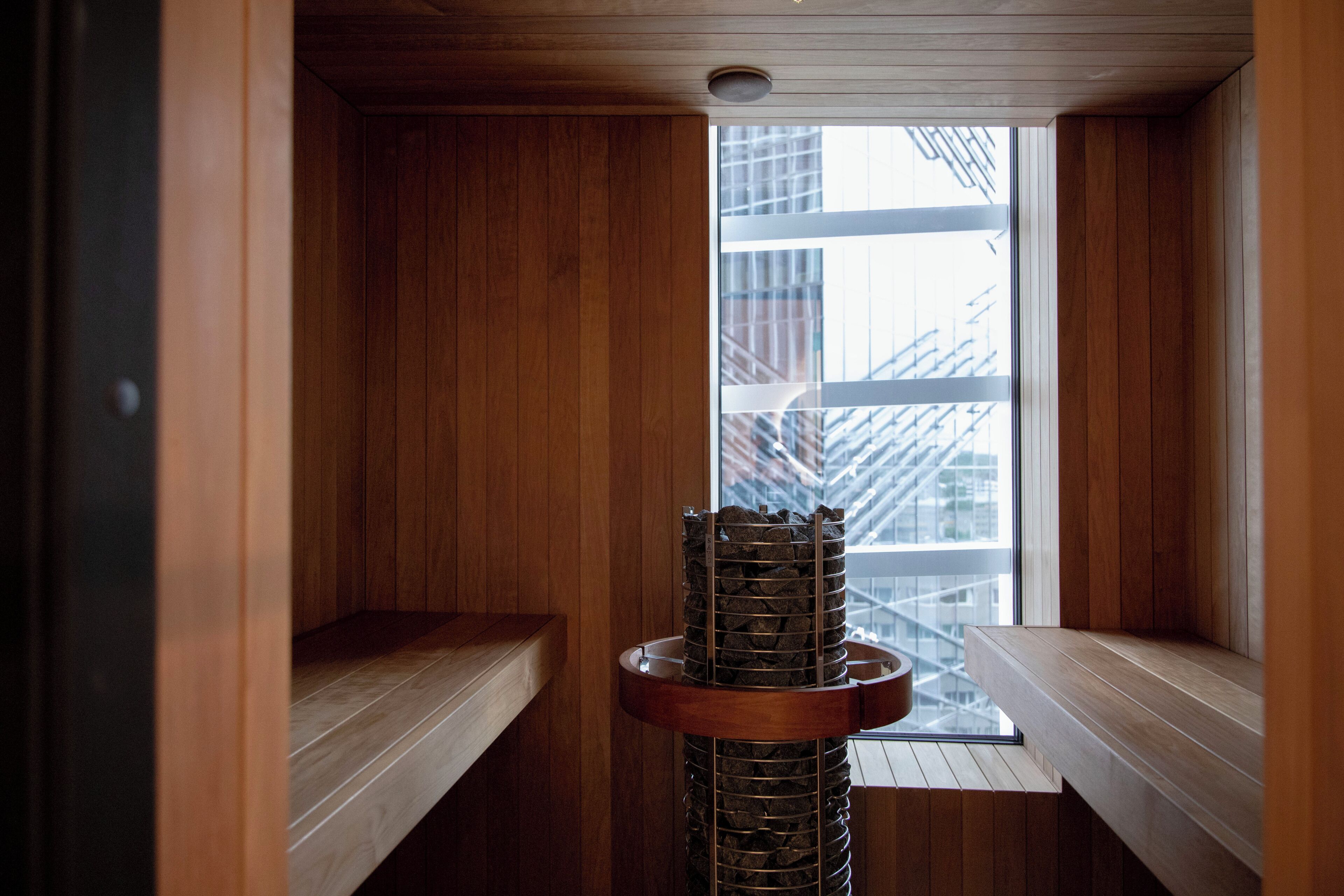 A sauna with wooden walls and benches, a central stone pillar, and a view of buildings through a window.