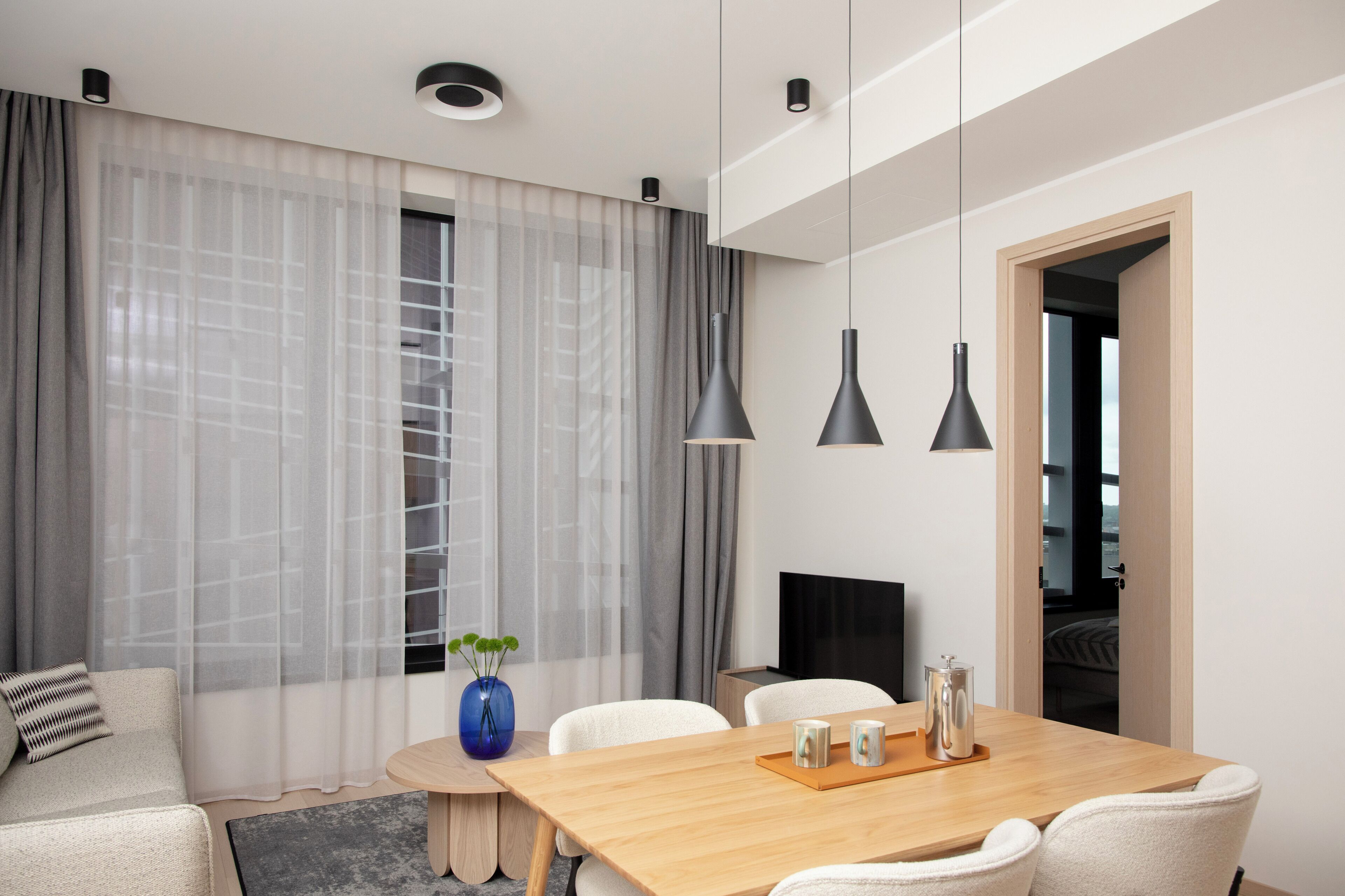 Une chambre d’hôtel moderne avec une table à manger en bois, des chaises blanches, un téléviseur et une grande fenêtre ornée de rideaux légers.
