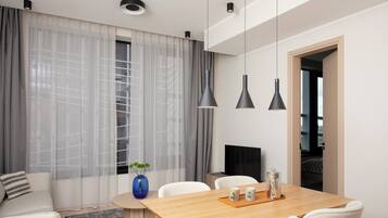 Une chambre d’hôtel moderne avec une table à manger en bois, des chaises blanches, un téléviseur et une grande fenêtre ornée de rideaux légers.