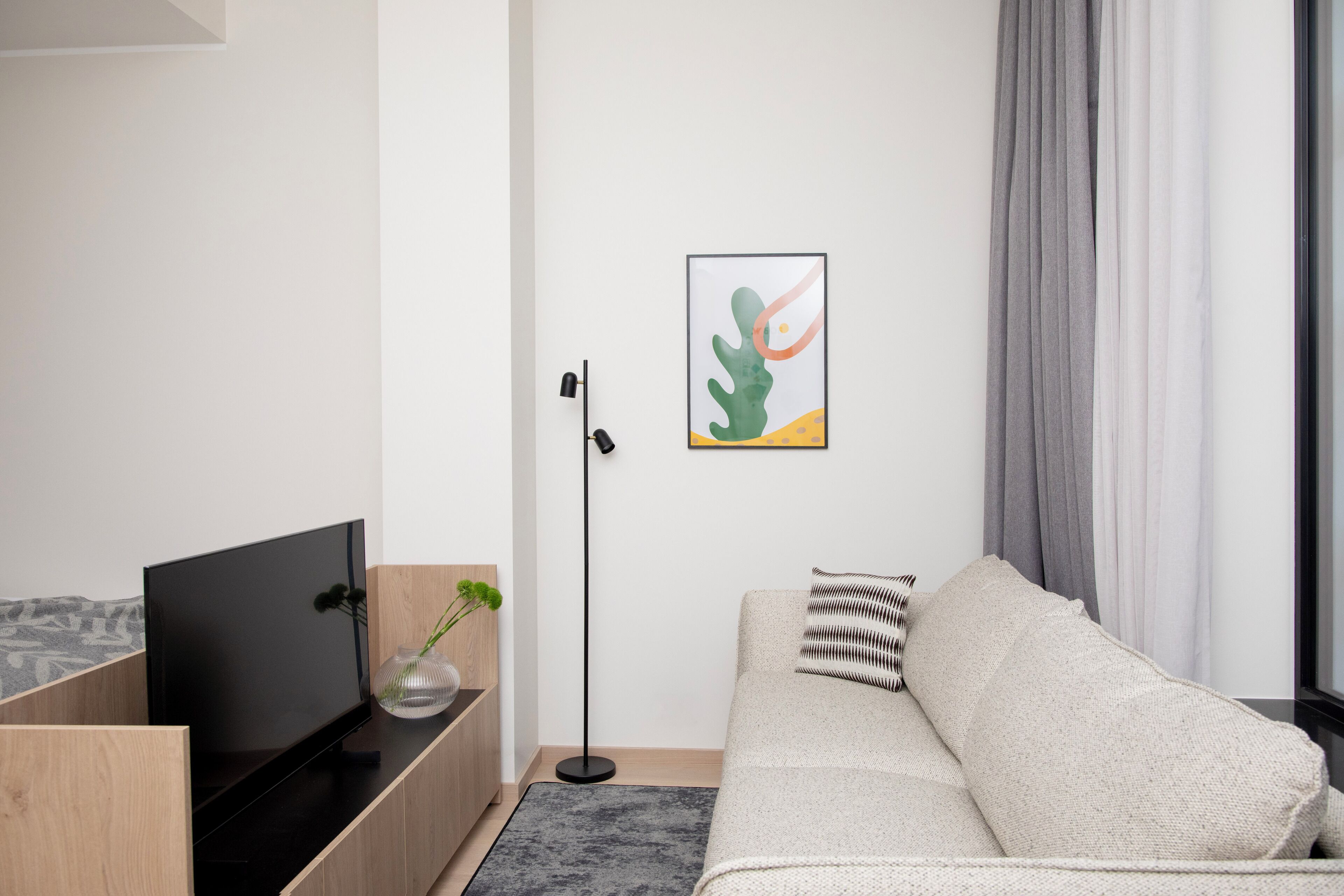 Una sala de estar moderna con un televisor de pantalla plana, una mesita de madera, un jarrón con plantas verdes, un sofá gris con un cojín a rayas y una obra de arte abstracto enmarcada en la pared.