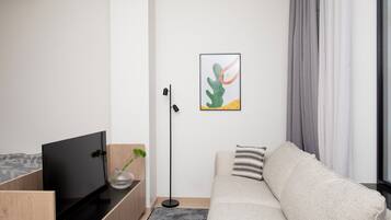 A modern living room with a flat-screen TV, a wooden side table, a vase with green plants, a gray sofa with a striped pillow, and a framed abstract art piece on the wall.