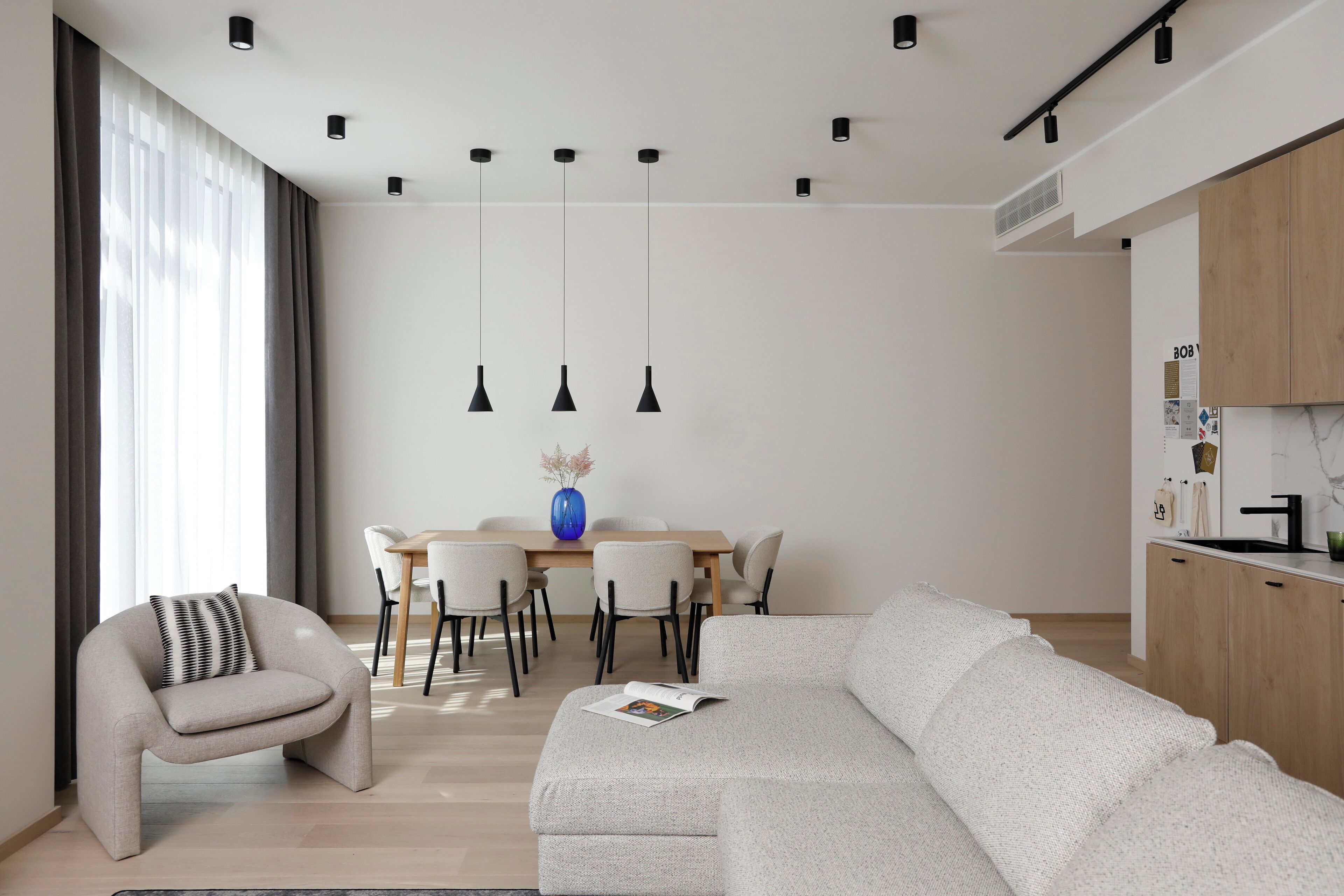 A modern living room with a sofa, a chair, a dining table with chairs, pendant lights, and a kitchen area with cabinets and a sink.