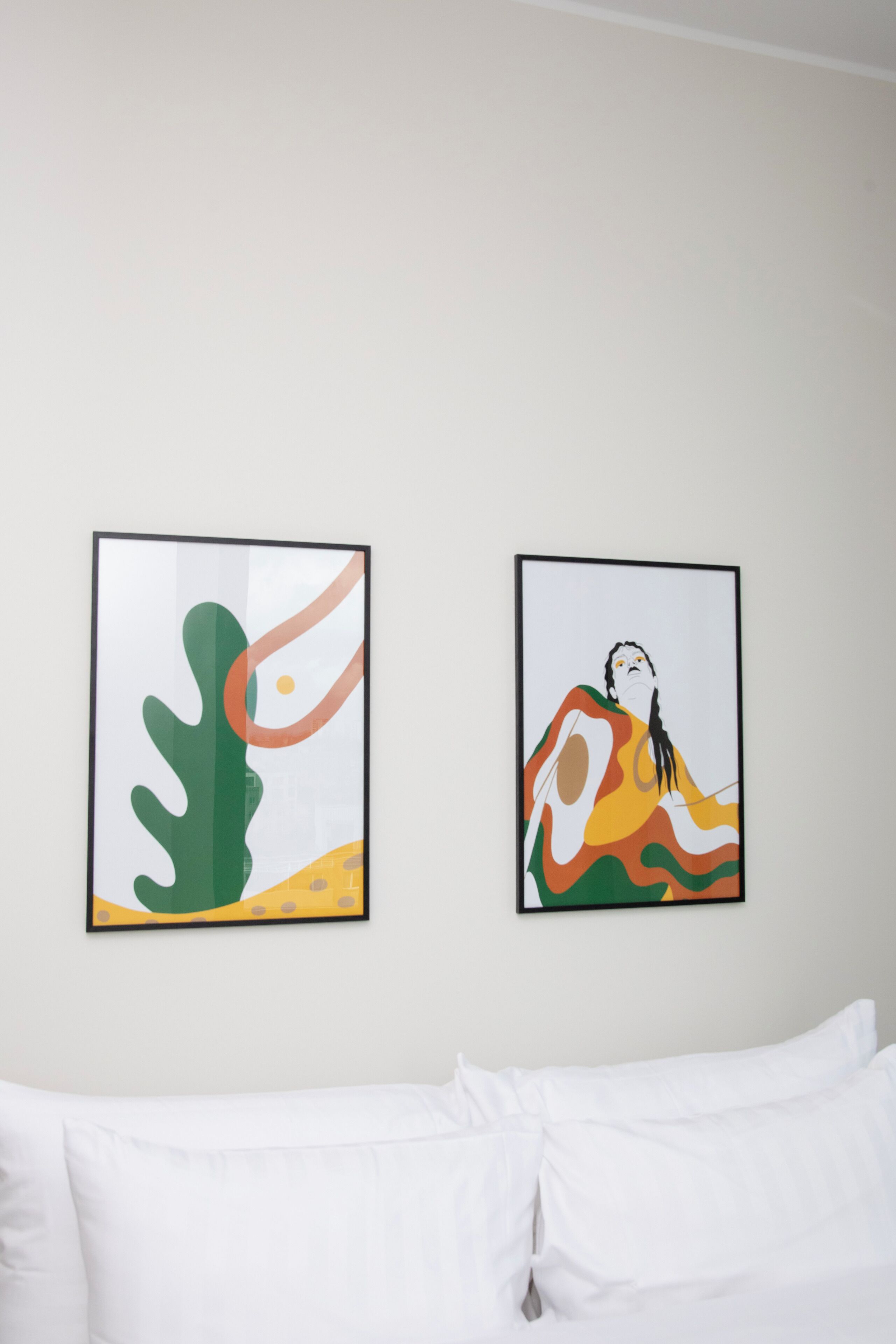 Two framed artworks on a plain wall above a white bed with white bedding.