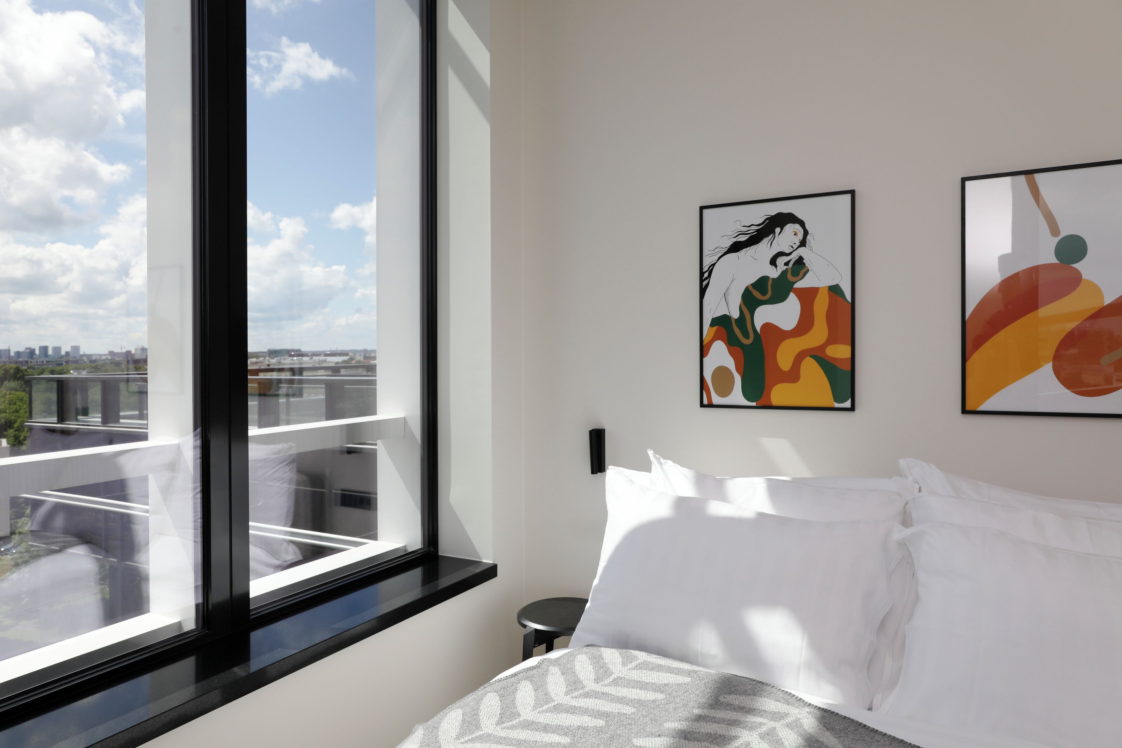 Habitación de hotel con un ventanal grande, una cama con ropa de cama blanca y dos cuadros abstractos en la pared.