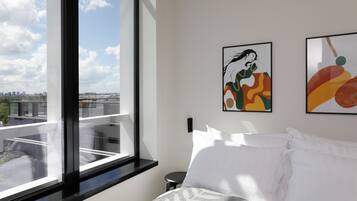 Habitación de hotel con un ventanal grande, una cama con ropa de cama blanca y dos cuadros abstractos en la pared.