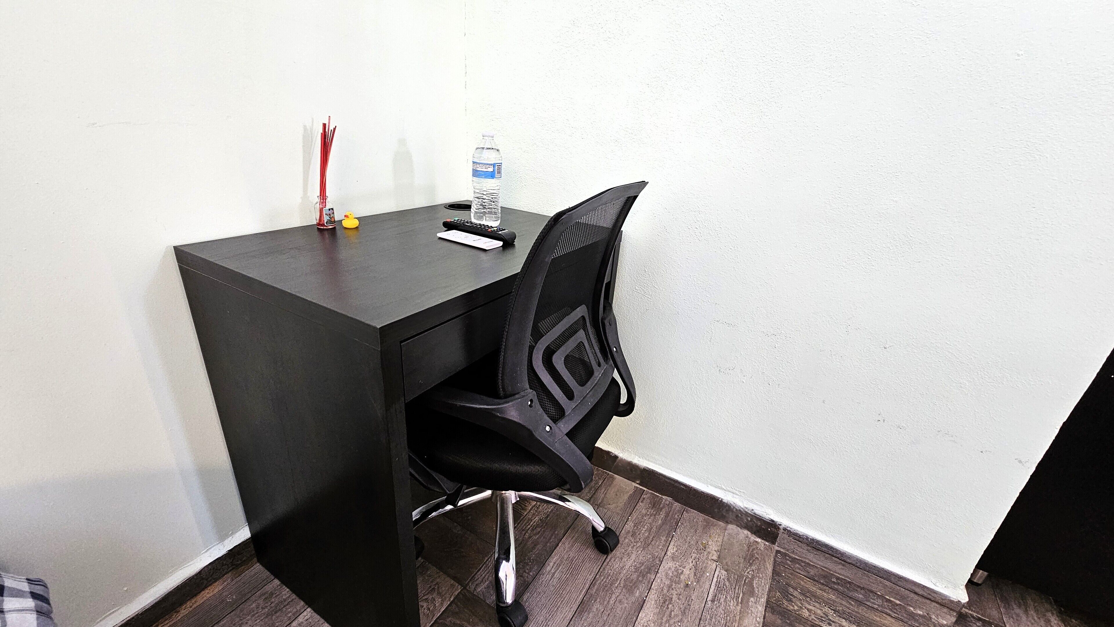 Een klein, donker houten bureau met een zwarte mesh bureaustoel. Op het bureau staan een waterfles en een rode pen.