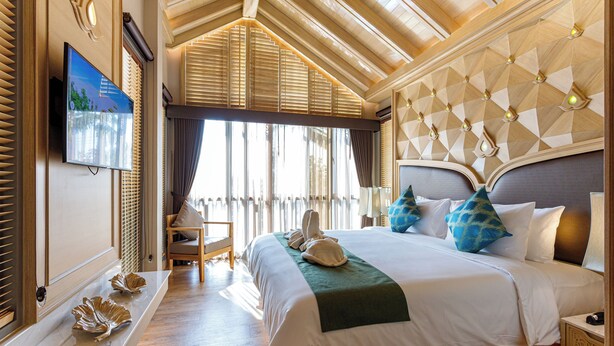 A spacious bedroom with a large bed, a television on the wall, and a view of the outdoors through large windows.