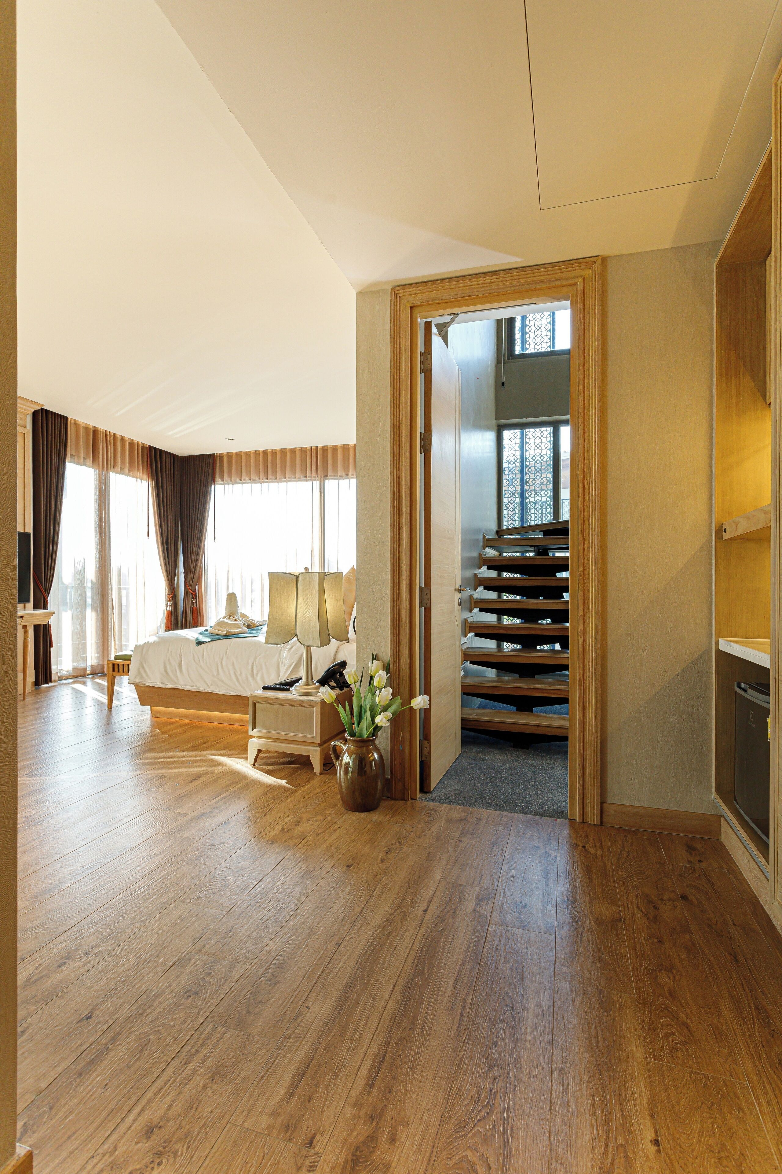 A hotel room with a wooden floor, a bed, a desk with a lamp, a vase with flowers, and a view of a staircase.