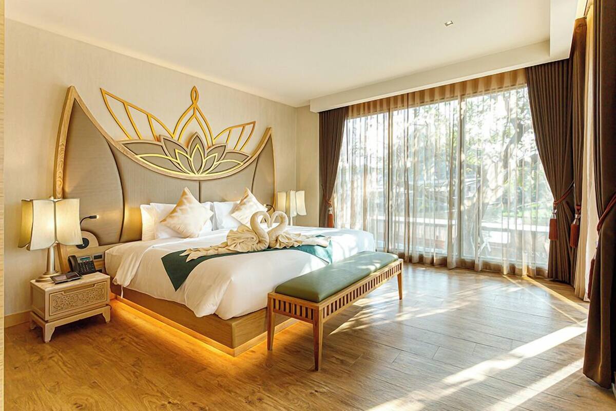 Une chambre d’hôtel spacieuse, dotée d’un grand lit, d’une table de chevet avec une lampe, d’un banc et offrant une vue sur les arbres par la fenêtre.