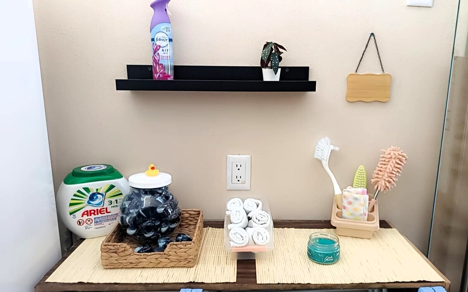A bathroom with a shelf holding a bottle of Febreze, a potted plant, and a wooden sign. There are toiletries on a tray, including a bottle of Ariel dish soap, a jar of blackheads, and a container of white towels.