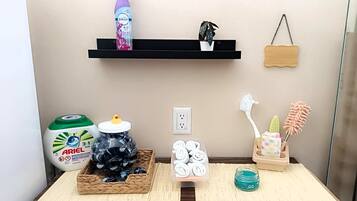 A bathroom with a shelf holding a bottle of Febreze, a potted plant, and a wooden sign. There are toiletries on a tray, including a bottle of Ariel dish soap, a jar of blackheads, and a container of white towels.