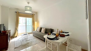 A modern living room with a sofa, dining table, and a view of the outside. - QUARTEIRA CENTRAL BEACH 1 by HOMING (Quarteira)