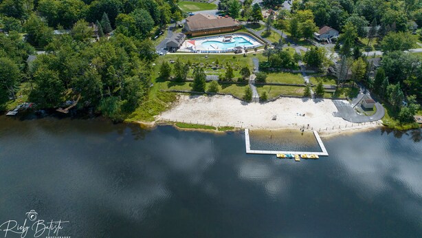 An aerial view of a resort with a swimming pool, beach, and forested area.