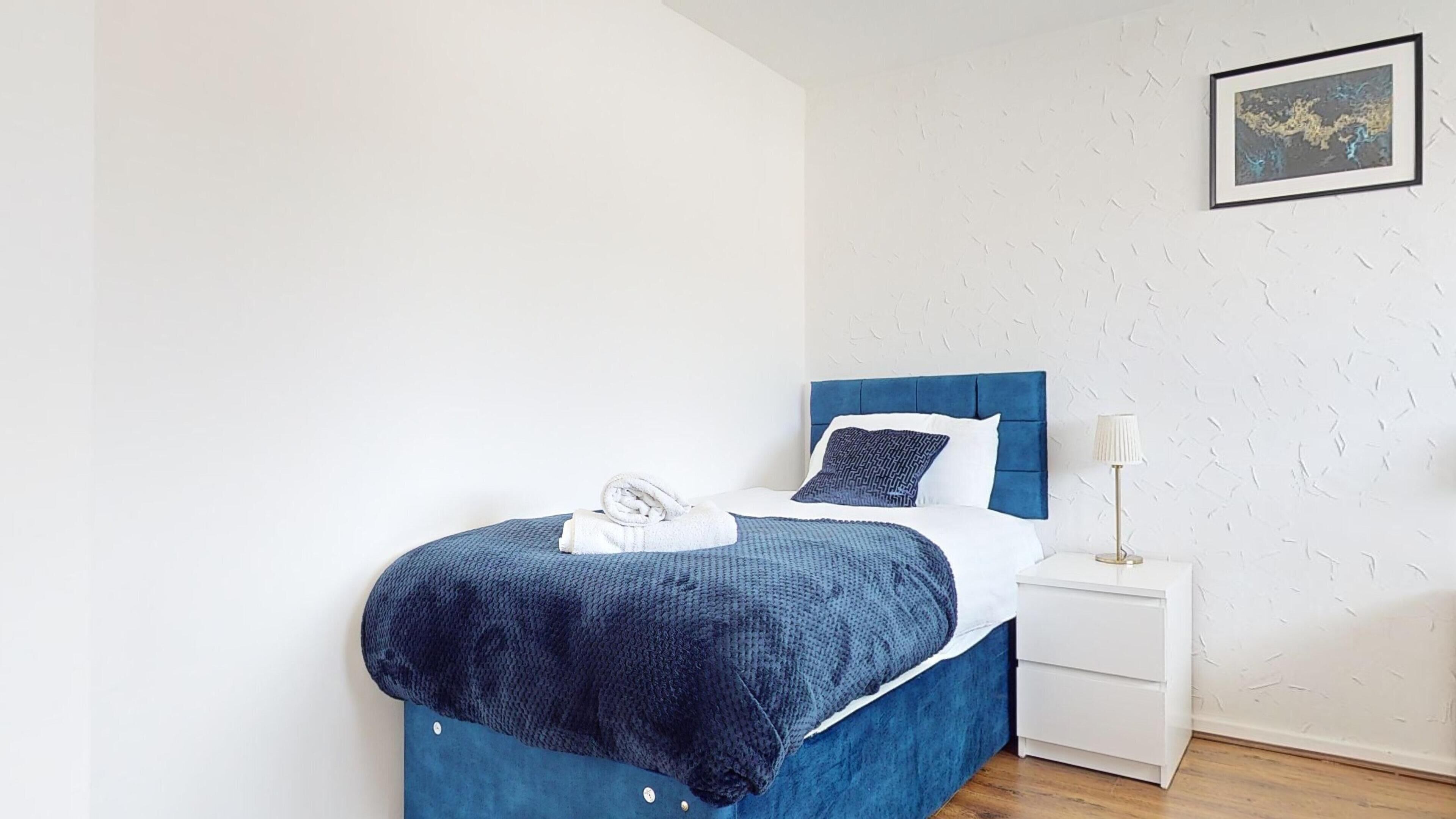 A neatly made bed with a blue headboard, a white nightstand with a lamp, and a textured white wall with a framed picture.