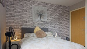 A bedroom with a brick-patterned wall, a bed with a leaf-patterned duvet, a nightstand with a fan, and a door. - Manchester Retreat - Modern 2Br Apartment (Manchester)