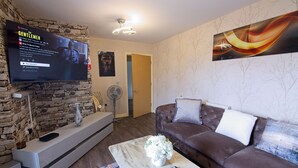 A modern living room with a stone accent wall, a flat-screen TV displaying 'The Gentleman', a brown leather sofa, and a marble coffee table. - Manchester Retreat - Modern 2Br Apartment (Manchester)