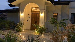 Una casa a due piani con un portale ad arco centrale, un patio piastrellato e un giardino con diverse piante e luci decorative.
