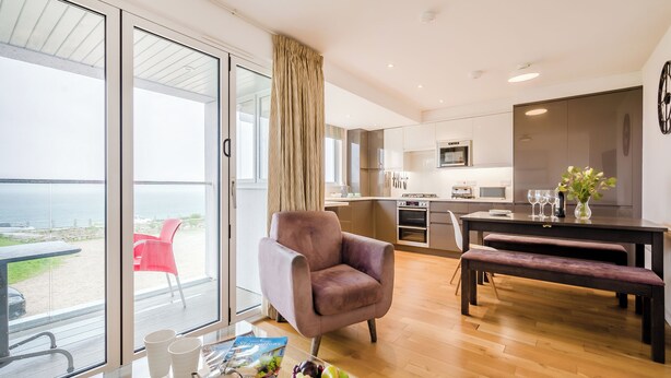 Ein modernes Wohnzimmer mit einem großen Glasfenster, das auf einen Balkon führt, einem Essbereich mit Tisch und Stühlen sowie einer Küche mit Edelstahlgeräten.
