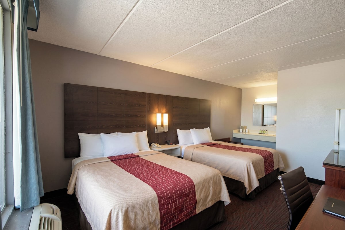 Une chambre d’hôtel avec deux lits, un bureau, une chaise, un miroir et une fenêtre avec des rideaux.