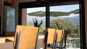 A dining area with wooden chairs and a table, large windows with mountain views, and outdoor umbrellas. - Appartement T3 bas de Villa (Vero)