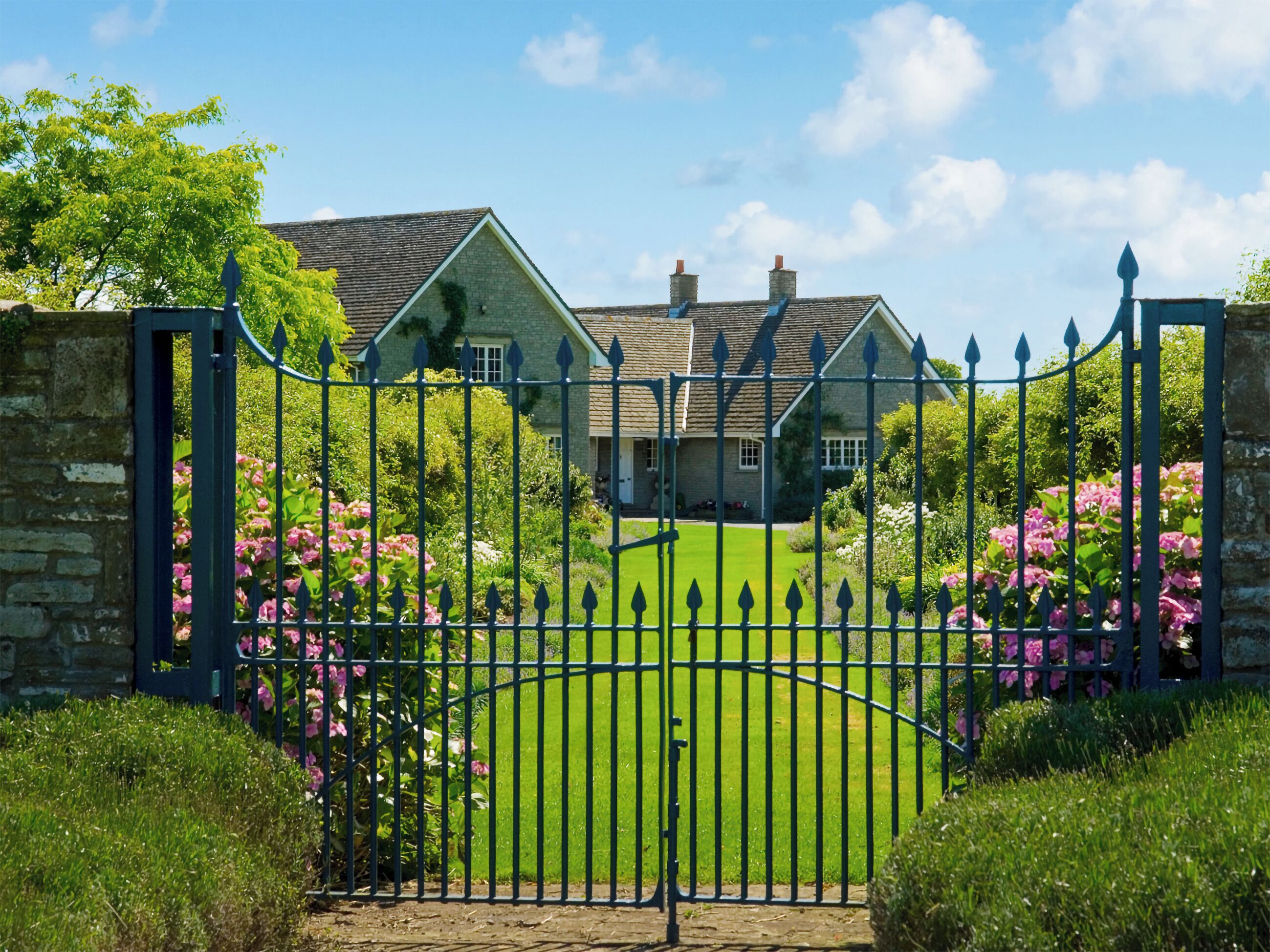 A gated entrance with a stone wall and a metal gate leading to a house with a green lawn and pink flowers.