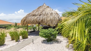 A thatched-roof structure with a paved walkway leading to it, surrounded by tropical plants and a clear sky.