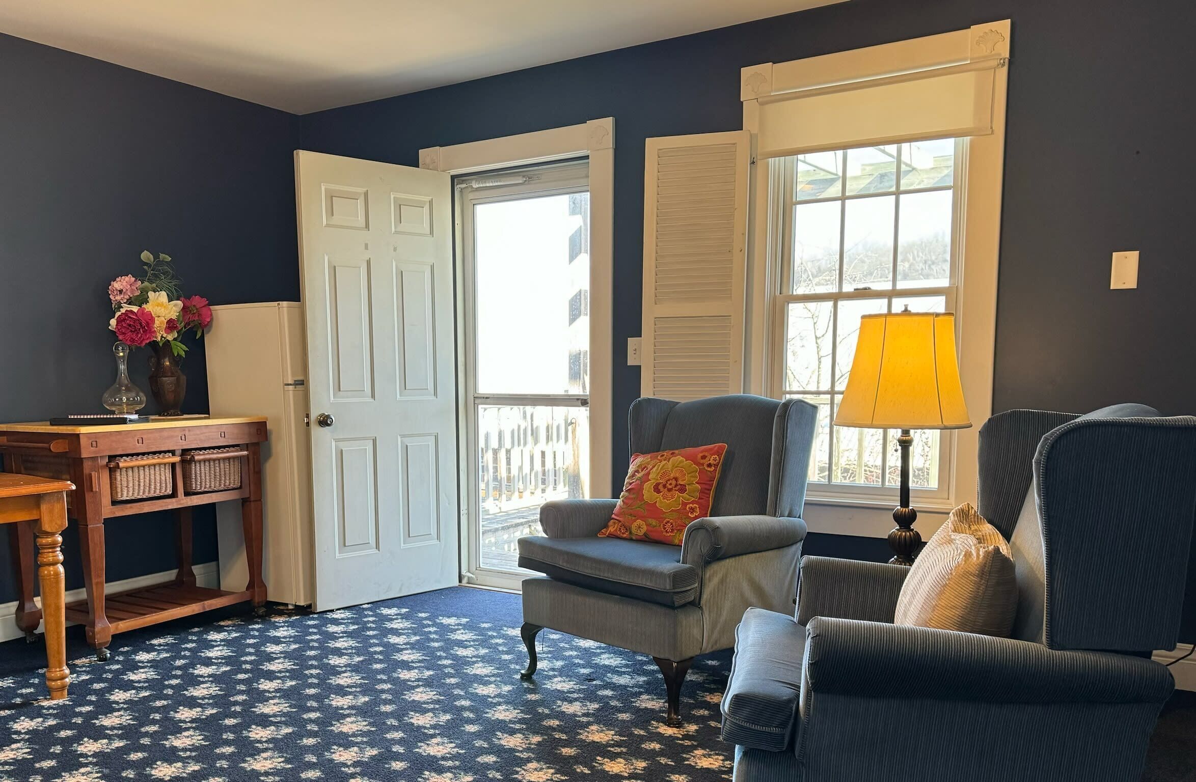 A room with a dark blue wall, a white door, a window with shutters, a grey armchair with a red pillow, a wooden table with baskets, and a blue patterned carpet.