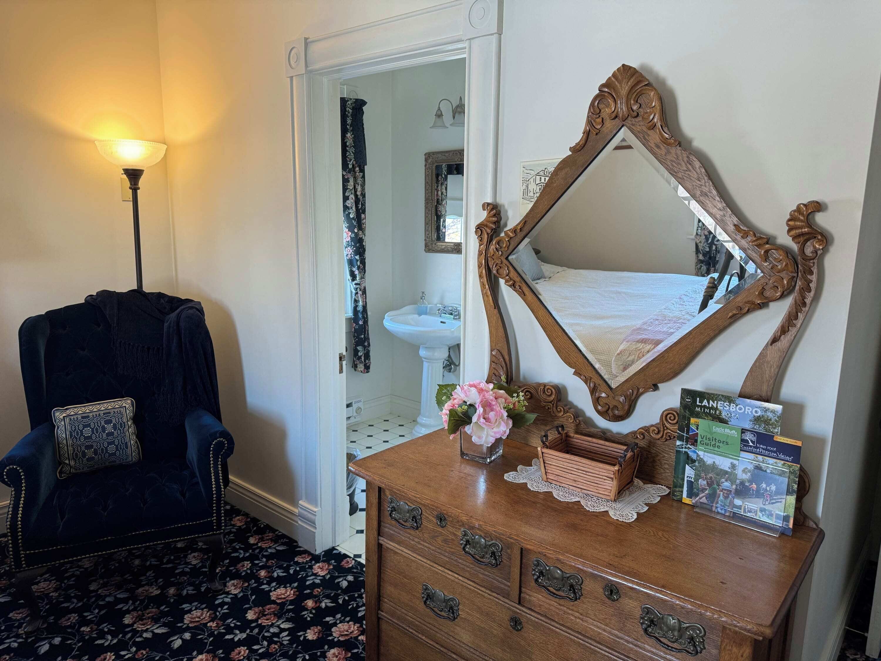 Ein Zimmer mit einem dunkelblauen Sessel, einer Holzkommode mit Schubladen, einem großen, verzierten Spiegel, einer Vase mit Blumen und einer Broschüre auf der Kommode.