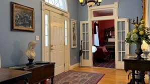 Un pasillo con suelo de madera, un candelabro, un busto y una puerta que da a otra habitación.