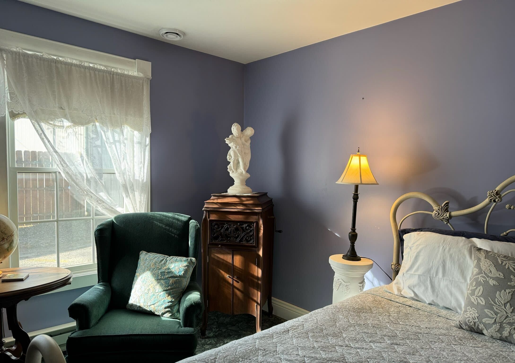 A bedroom with a bed, a green armchair, a small table with a globe, and a lamp.