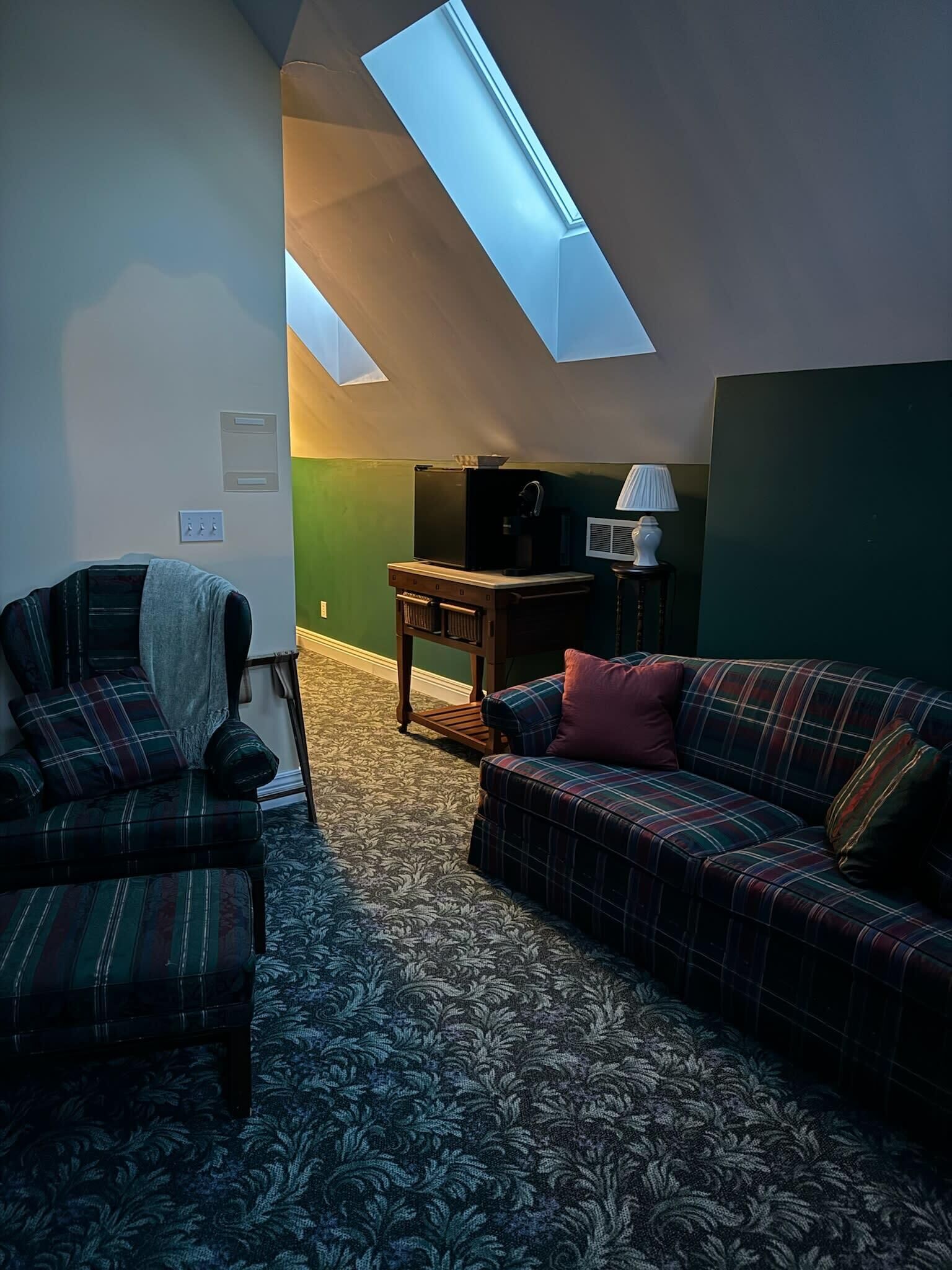 Una sala de estar acogedora con un sofá, un sillón y una mesita de centro. Un tragaluz permite el ingreso de luz natural, y una lámpara está sobre la mesa.