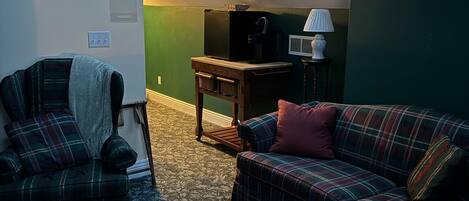 Una sala de estar acogedora con un sofá, un sillón y una mesita de centro. Un tragaluz permite el ingreso de luz natural, y una lámpara está sobre la mesa.