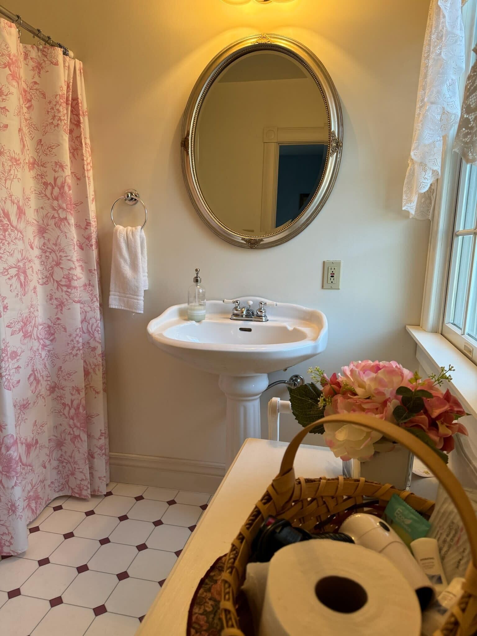 Une salle de bain avec un lavabo blanc, un miroir rond, un rideau de douche à motifs floraux et une fenêtre ornée de rideaux en dentelle.