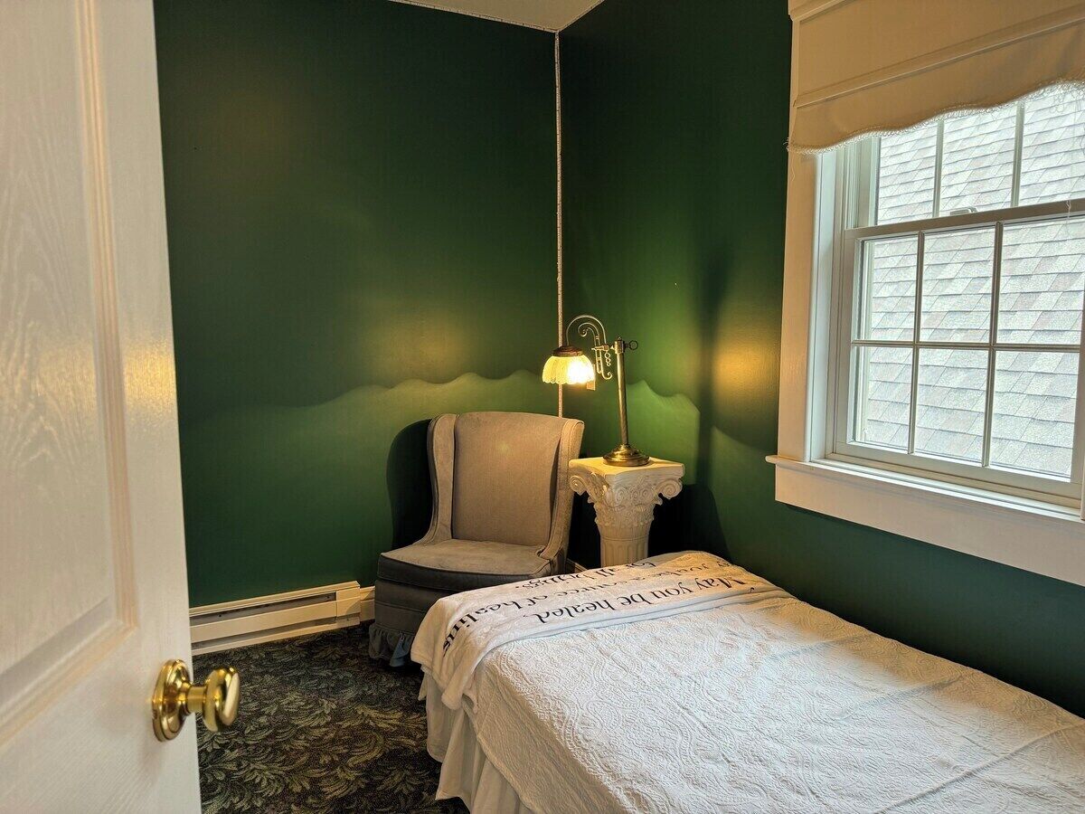 A small, cozy room with a single bed, a chair, a nightstand with a lamp, and a window with blinds.