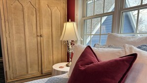 Un dormitorio con un armario de madera, una mesita de noche con lámpara y una cama con ropa de cama blanca y almohada roja.