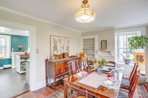 A dining area with a wooden table, chairs, and a chandelier. There is a window with a plant on the right and a door leading to another room on the left.