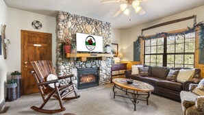 A cozy living room with a stone fireplace, a wooden rocking chair, a leather sofa, and a ceiling fan. - 2BD Cabin, Sleeps 6, near Hwy 76, Tanger Outlets- DISCOUNT TICKETS! (Branson)